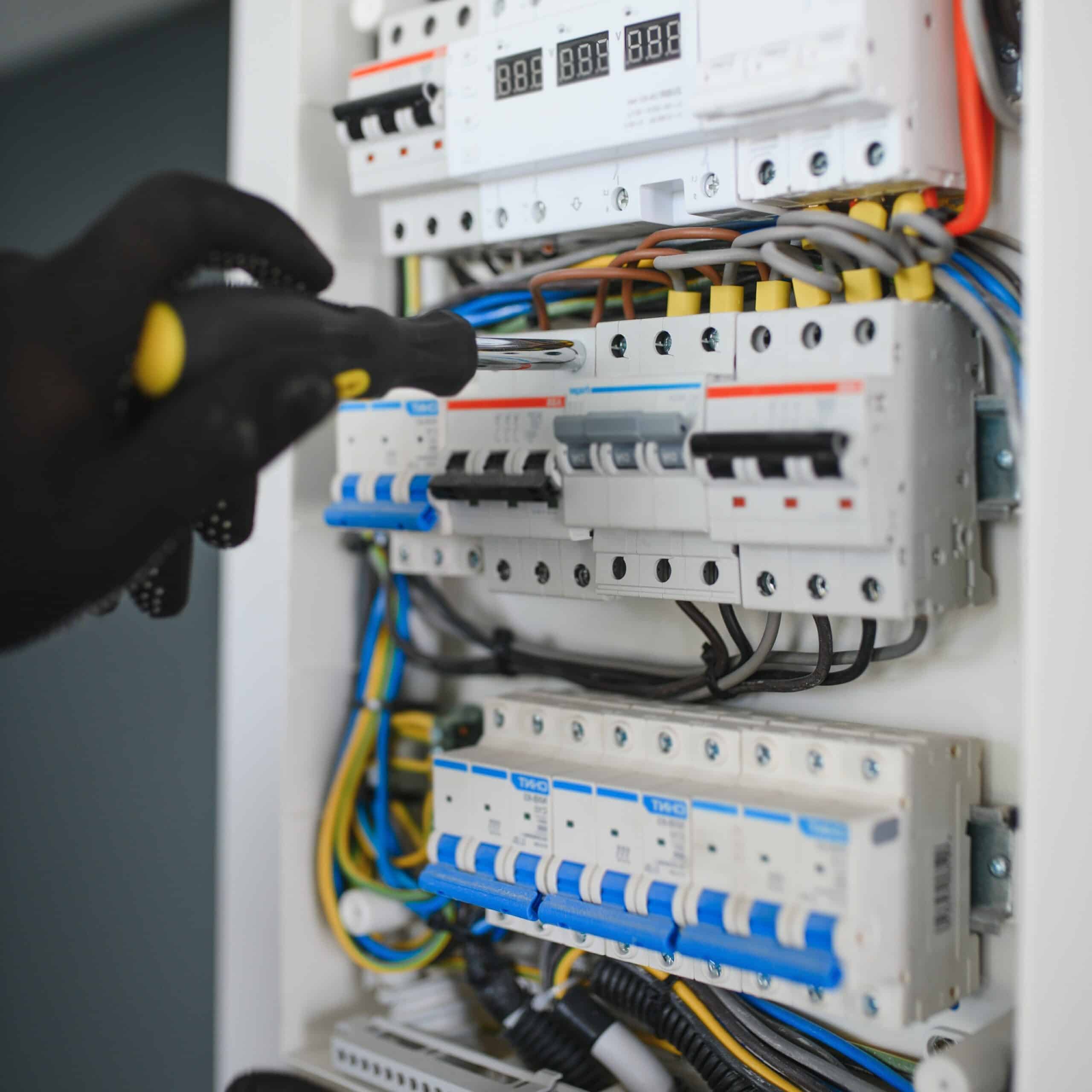 Électricien ganté serrant une vis sur un disjoncteur dans un tableau électrique moderne avec câblage apparent.