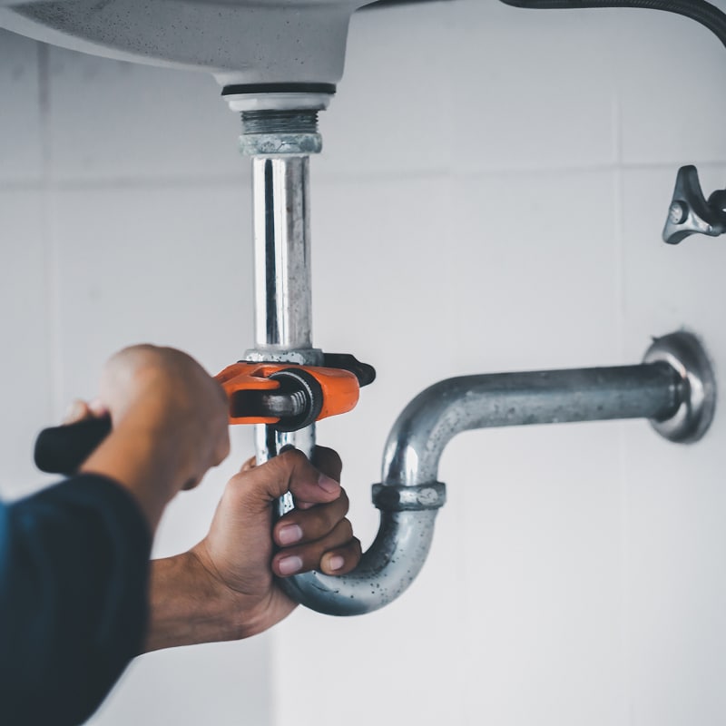 Plombier réparant une fuite d'eau sous un évier Mains d'un plombier serrant un tuyau d'évacuation sous un évier avec une clé à molette orange.