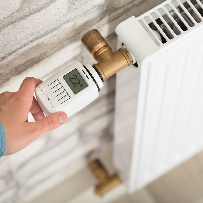 Thermostat Radiateur : Optimisez vos Économies d'Énergie Main réglant un thermostat numérique affichant 22°C sur un radiateur blanc pour optimiser la température.